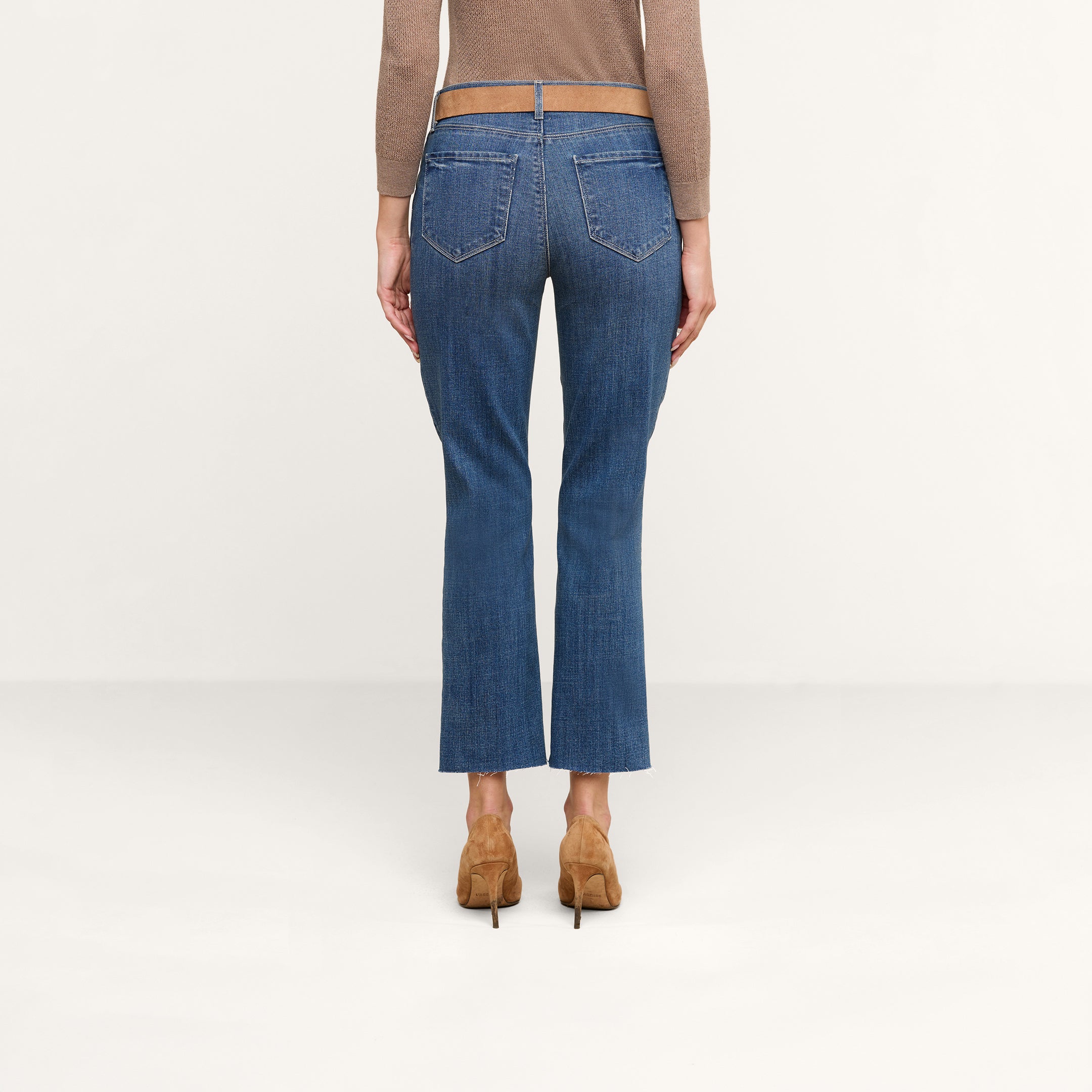 A person is shown from the back standing against a plain light background, wearing the Sada Cropped Slim-Leg Jean in denim blue with a raw hem, paired with a brown long-sleeve top and brown heeled shoes.