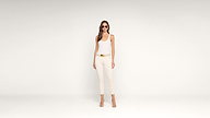 A woman stands against a plain white background wearing a white tank top, cream pants, beige heels, a tan belt with a gold buckle, and sunglasses. She has long brown hair and a neutral expression.