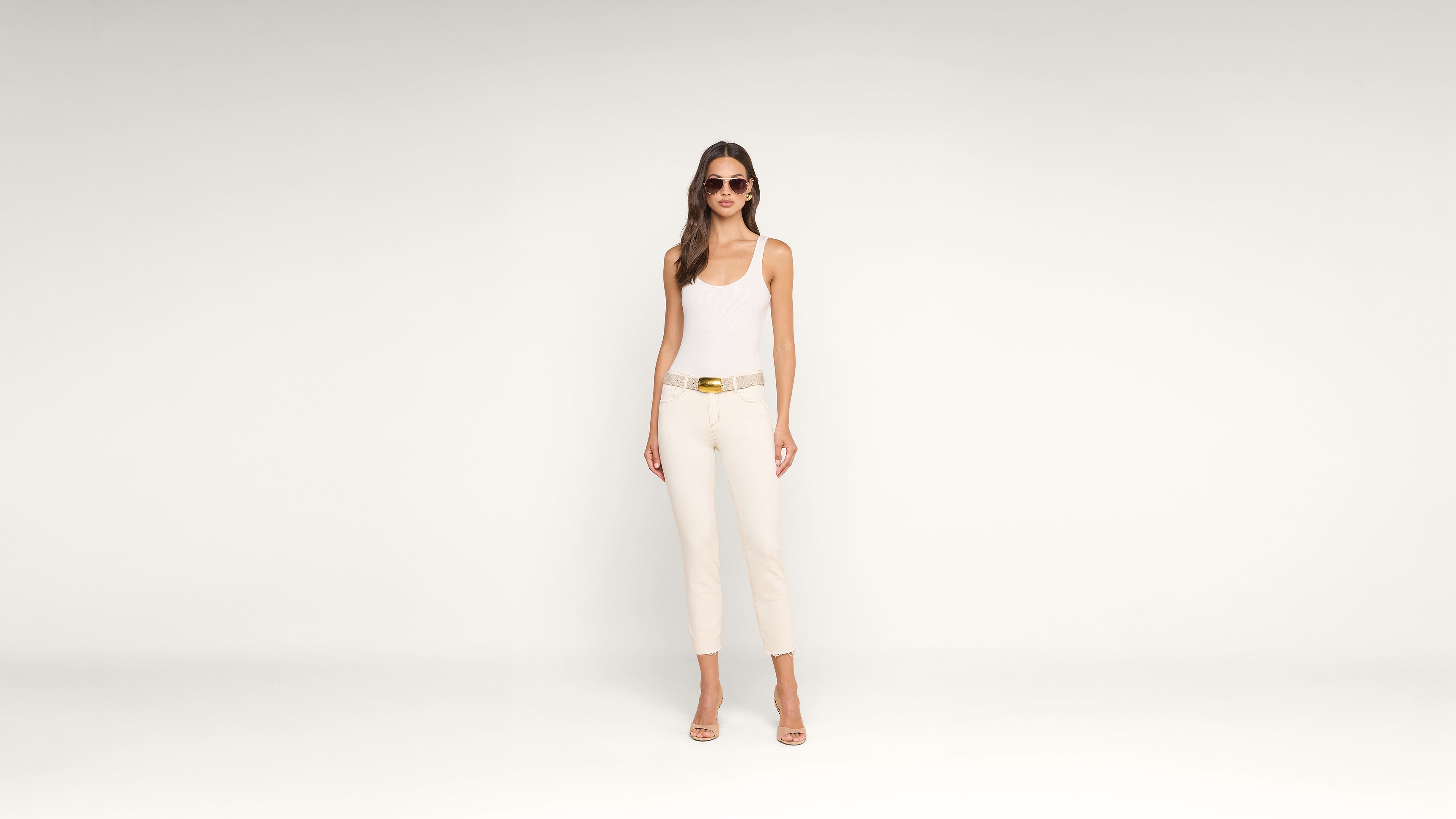 A woman stands against a plain white background wearing a white tank top, cream pants, beige heels, a tan belt with a gold buckle, and sunglasses. She has long brown hair and a neutral expression.