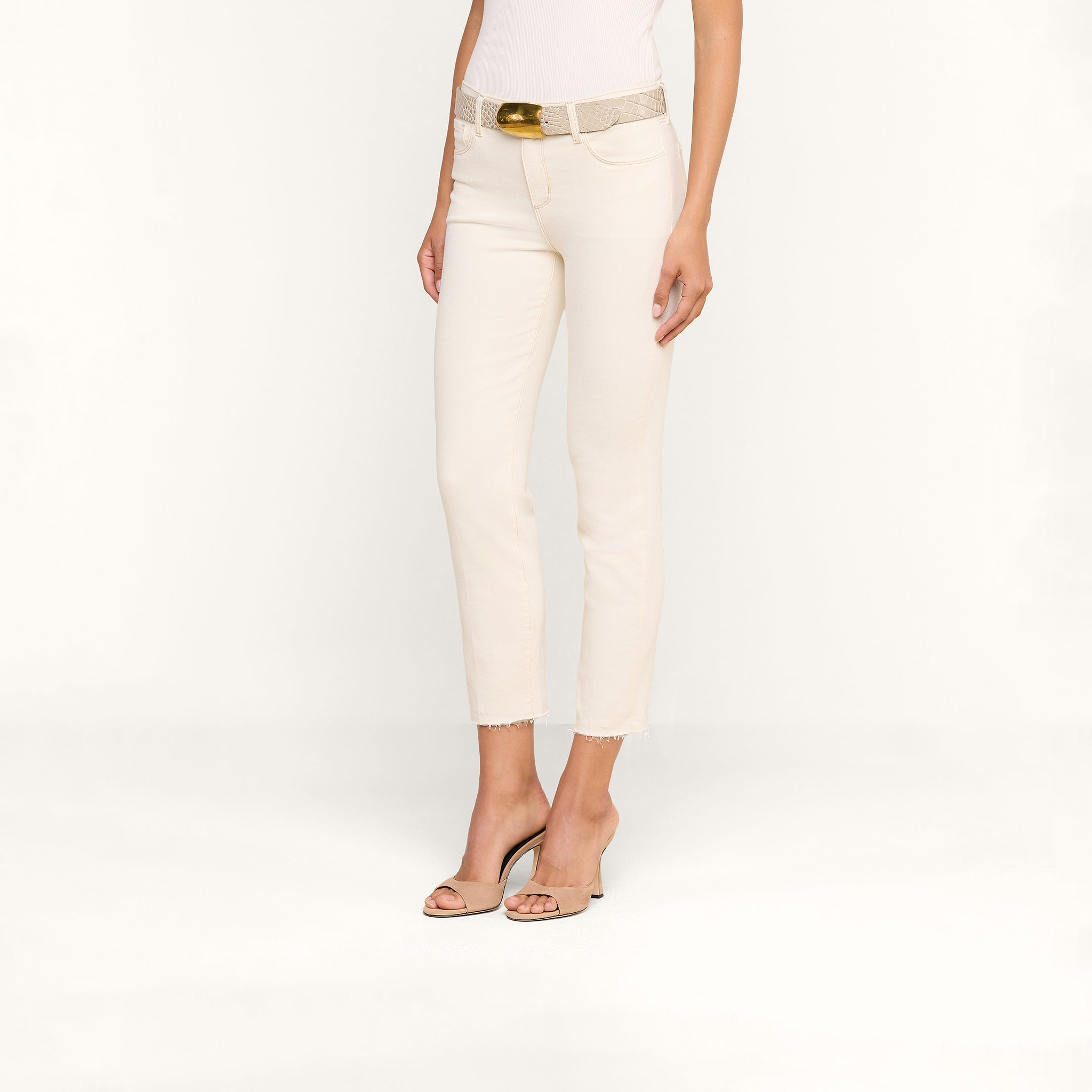 Une femme se tient debout devant un fond beige. Elle porte le jean court Sada Slim-Leg, confectionné dans un denim stretch haut de gamme blanc cassé, associé à un haut blanc, une ceinture beige texturée à boucle dorée et des escarpins nude à bout ouvert.