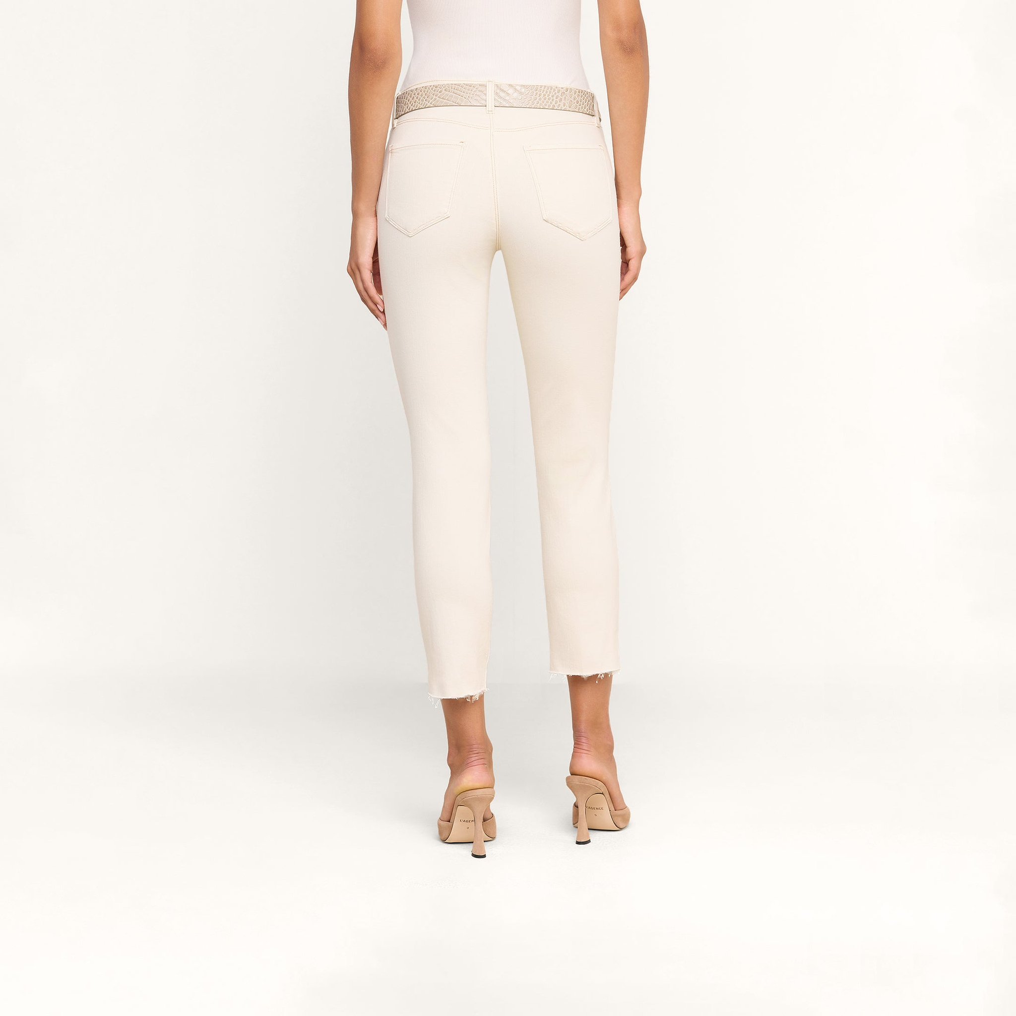 Une personne, de dos, porte un jean court Sada Slim-Leg aux ourlets effilochés, une ceinture texturée beige et des sandales à talons beiges, sur un fond beige uni.