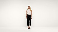 A woman stands facing forward against a plain white background, wearing a sleeveless white top, black pants, a black belt, and black heels.