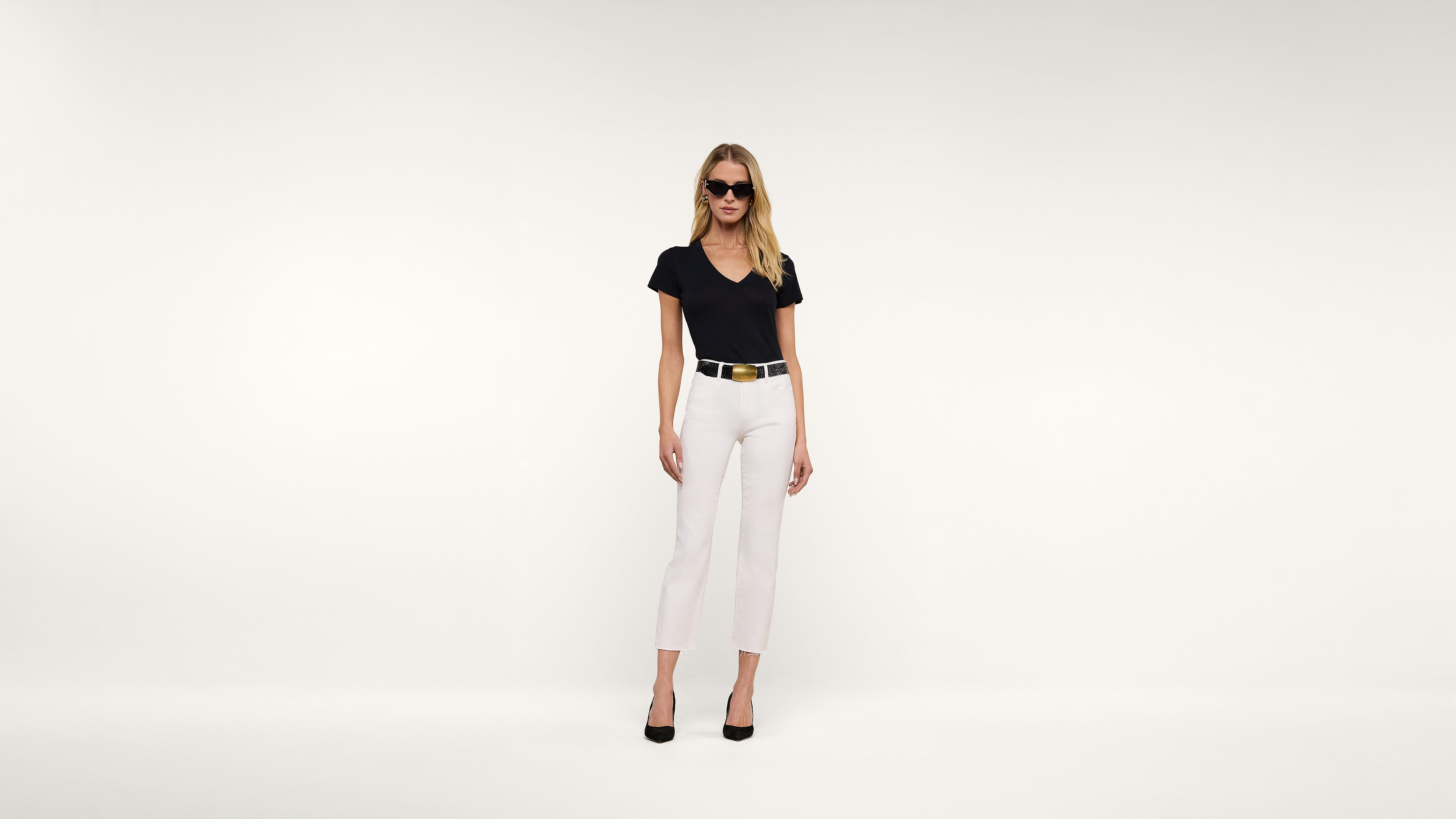 A woman in sunglasses, a black shirt, white pants, and black heels stands against a plain white background.