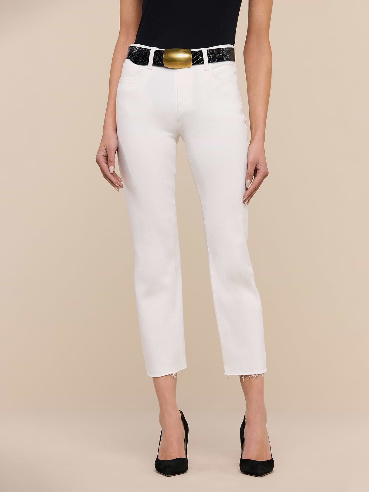 A person wearing Sada Slim-Leg Cropped Jeans, a black belt with a large gold buckle, a black sleeveless top, and black pointed-toe heels stands against a beige background.
