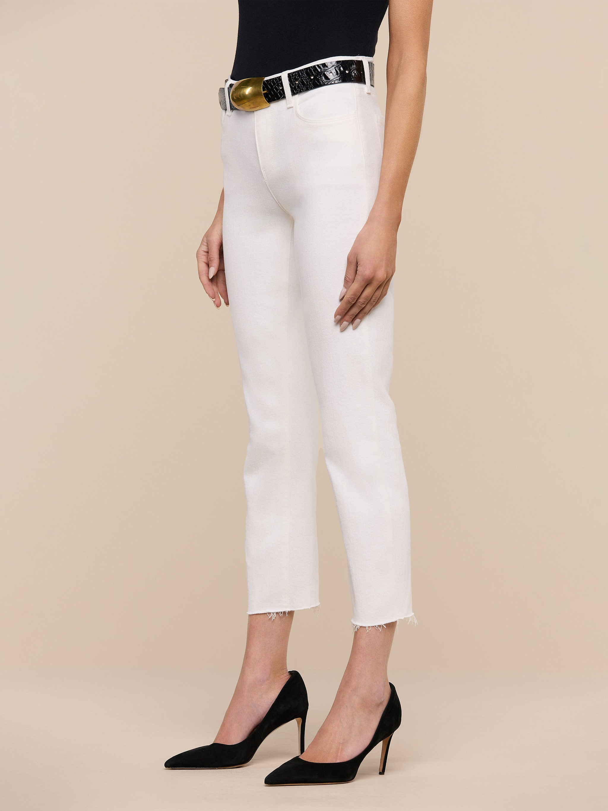 A woman stands against a white background wearing the Sada Slim-Leg Cropped Jean in stretch denim, paired with a black belt with a gold buckle, a black sleeveless top, and black high heels.