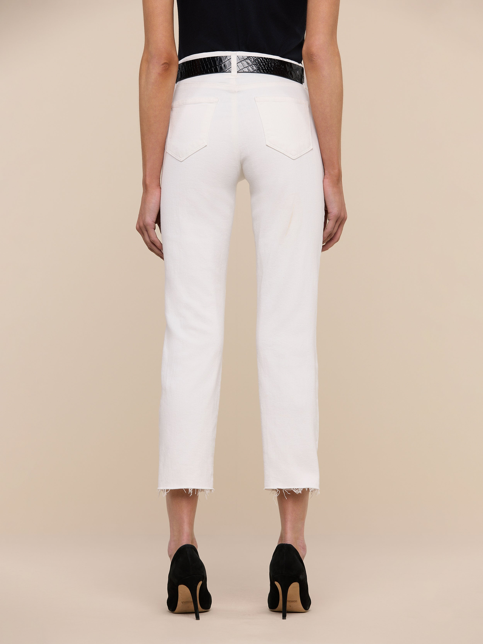A person stands facing away, wearing the Sada Slim-Leg Cropped Jean—a high-rise white jean with frayed hems—paired with a black belt, black heels, and a dark top, against a plain white background.