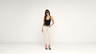 A woman wearing sunglasses, a black tank top, white pants, and black heels stands confidently in a minimalist white studio, looking at the camera.