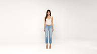 Woman standing against a plain white background, wearing a white tank top, light blue jeans, and beige high heels, looking directly at the camera with arms relaxed at her sides.
