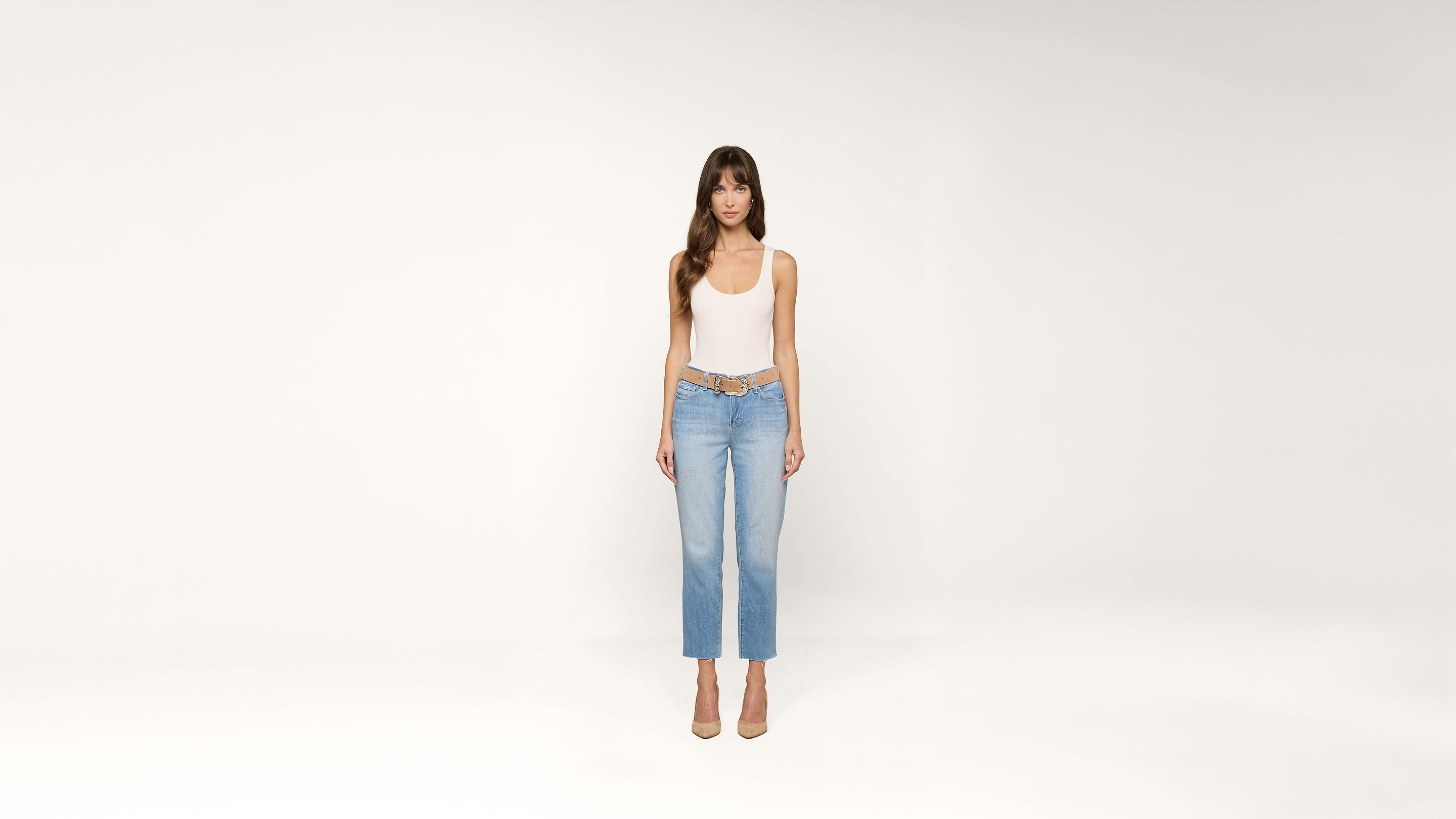 Woman standing against a plain white background, wearing a white tank top, light blue jeans, and beige high heels, looking directly at the camera with arms relaxed at her sides.
