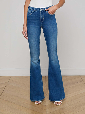 Wearing L'AGENCE Bell Flare Jean in high-rise blue stretch denim, a white sleeveless top, and white open-toe heels, the person stands on a wooden floor against a plain light wall with their head and upper body out of frame.