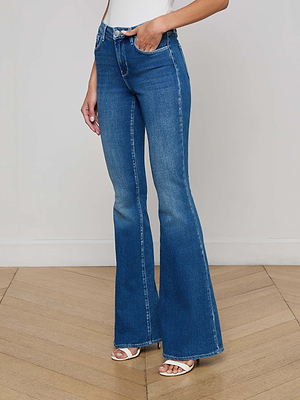 A person models the L'AGENCE Bell Flare Jean—high-rise, blue stretch denim—paired with white heeled sandals. They stand on a light wooden floor against a plain wall. The image is cropped at the shoulders.