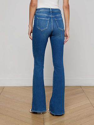 Wearing the L'AGENCE Bell Flare Jean in blue stretch denim and a white top, a person stands on a wooden floor, photographed from the back against a plain light wall.