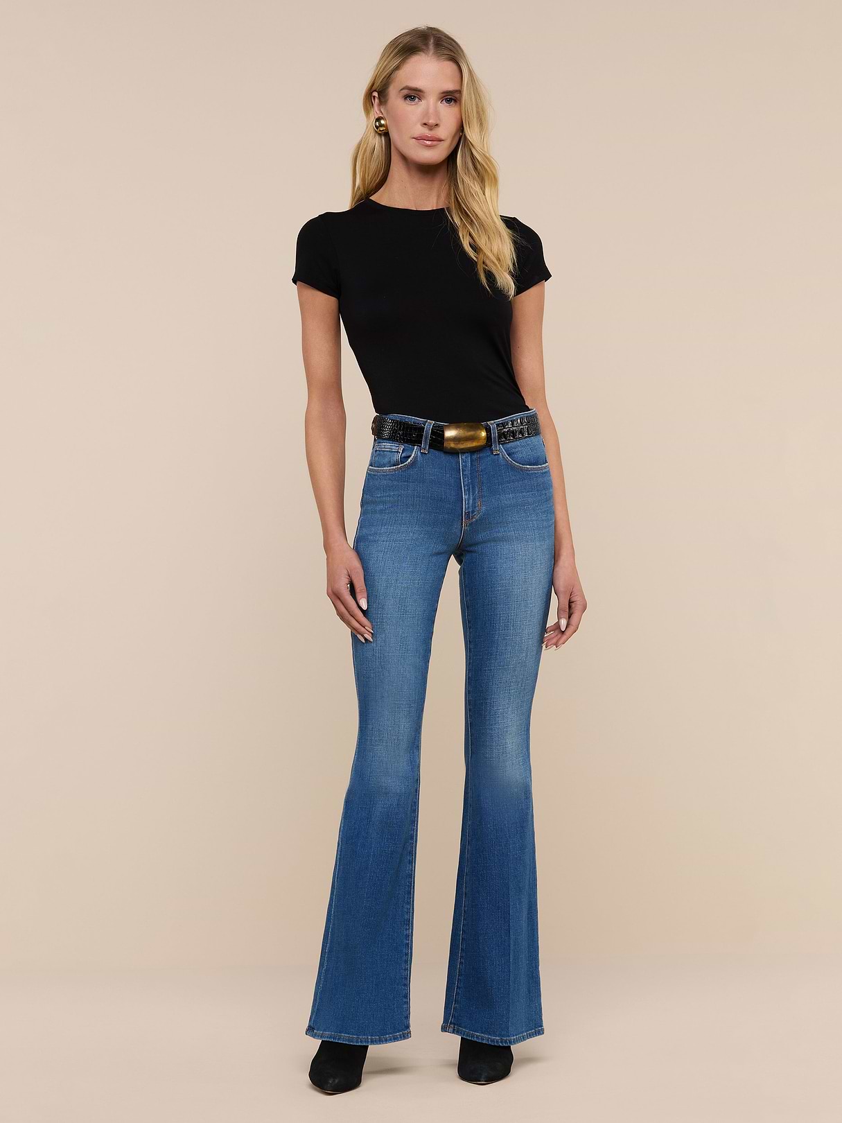 A woman stands facing forward against a plain white background, wearing a black short-sleeve top, the Bell Flare Jean in blue vintage-inspired premium stretch denim, a black belt with a gold buckle, and black shoes.