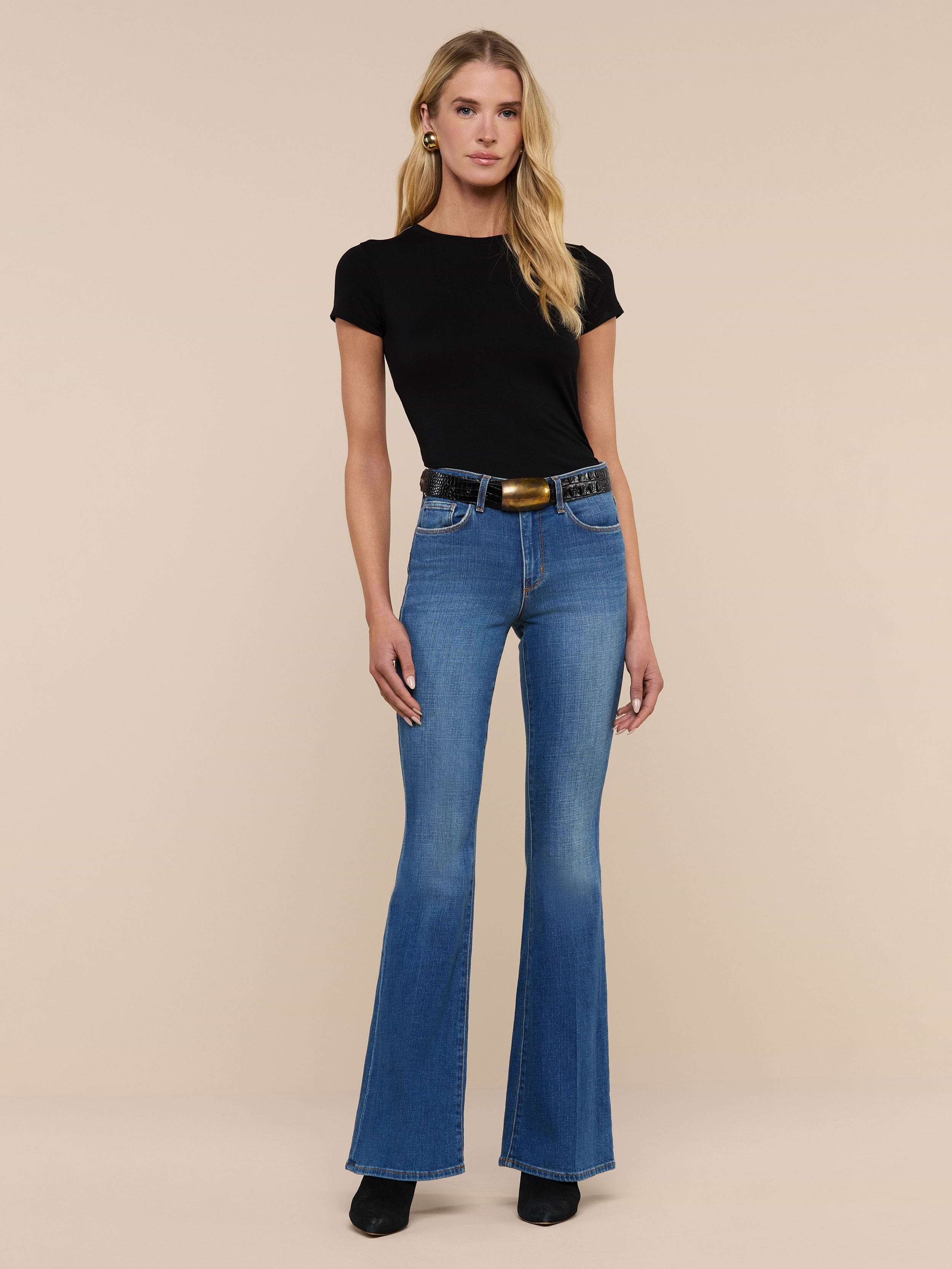 A woman stands facing forward against a plain white background, wearing a black short-sleeve top, the Bell Flare Jean in blue vintage-inspired premium stretch denim, a black belt with a gold buckle, and black shoes.