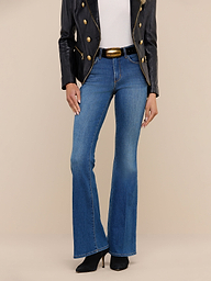 A person wearing the Bell Flare Jean—a pair of blue, vintage-inspired bell-bottom jeans crafted from premium stretch denim—paired with a black jacket, white top, and black heeled boots stands against a plain white background.