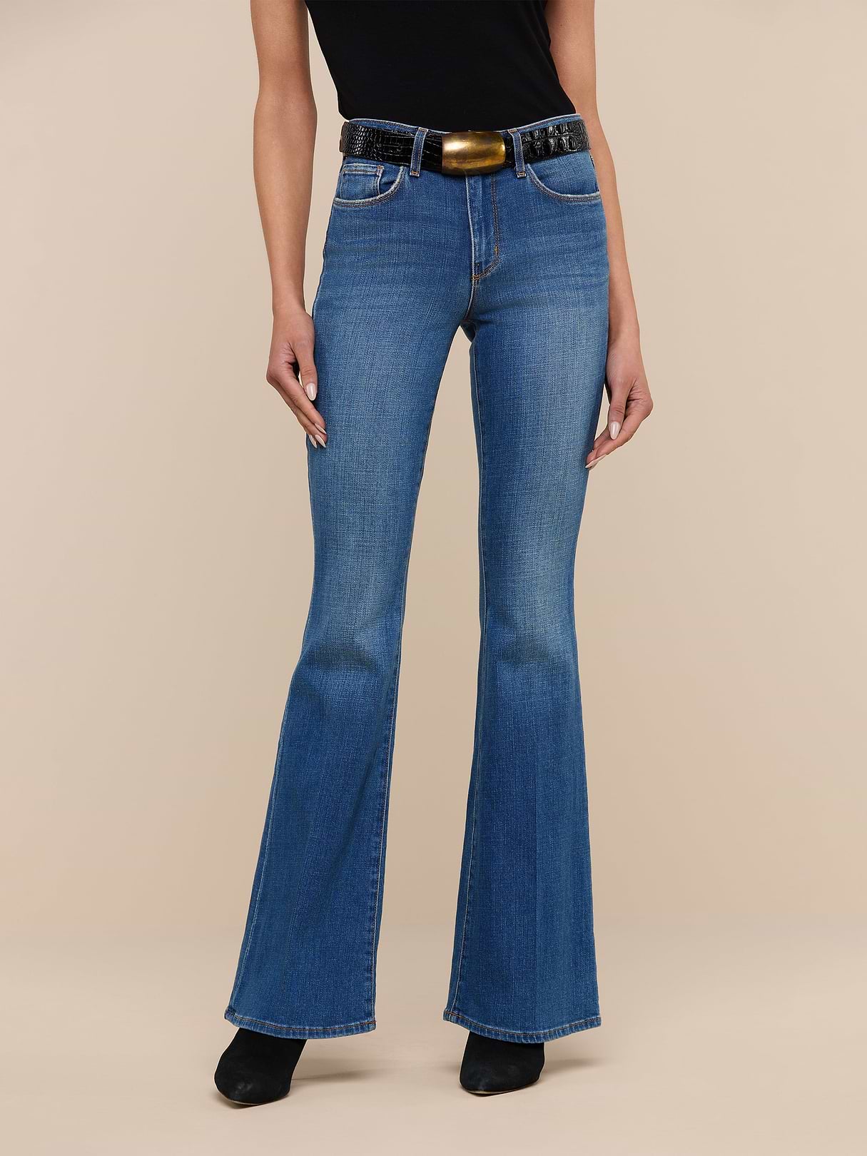 A person wearing the Bell Flare Jean in vintage-inspired blue premium stretch denim, styled with a black sleeveless top, a wide black belt featuring a large buckle, and black shoes, stands against a plain white background.