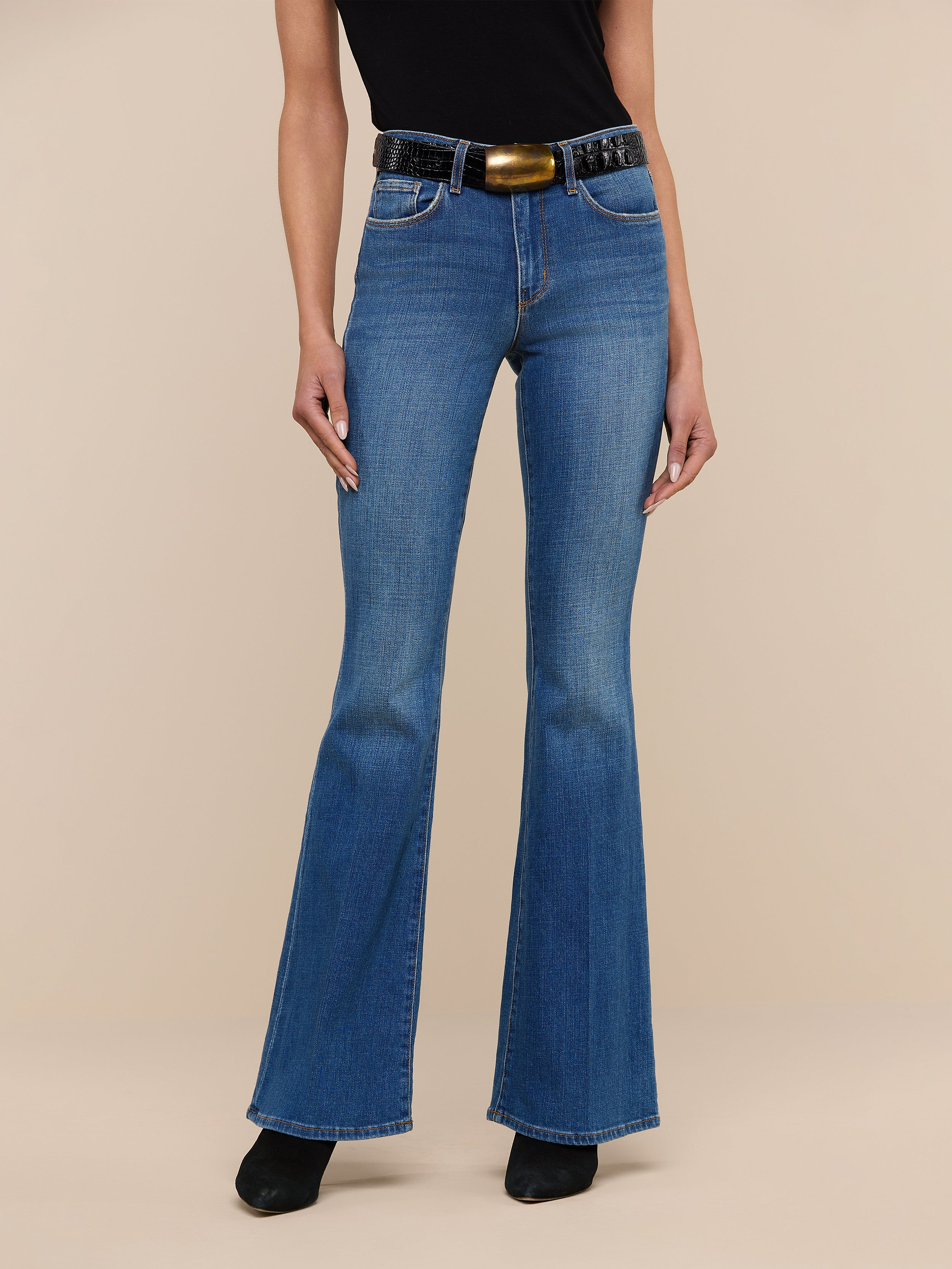 A person wearing the Bell Flare Jean in vintage-inspired blue premium stretch denim, styled with a black sleeveless top, a wide black belt featuring a large buckle, and black shoes, stands against a plain white background.