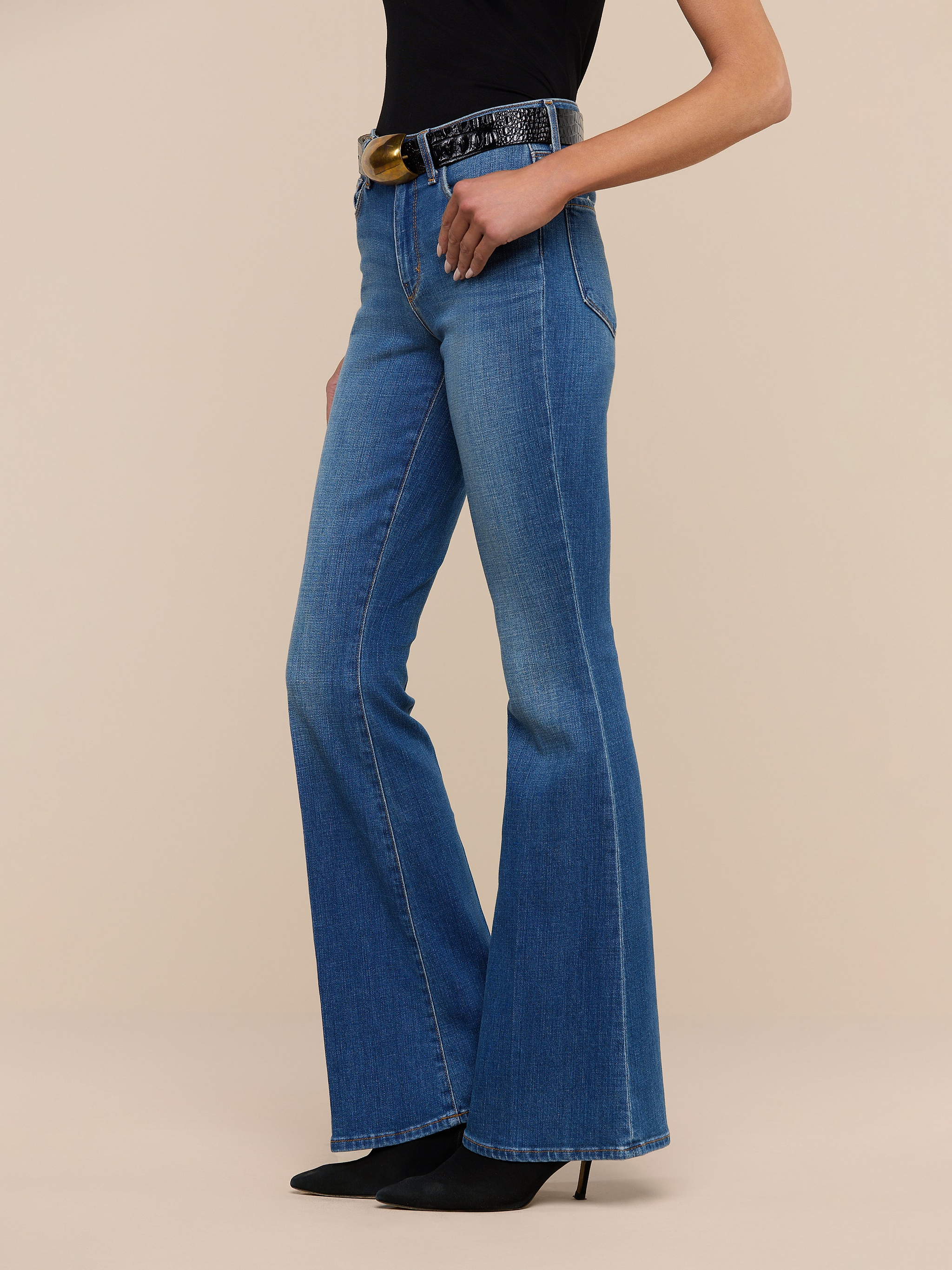 A woman stands against a plain white background wearing the Bell Flare Jean, vintage-inspired blue bell-bottom jeans crafted from premium stretch denim, paired with black pointed-toe heels, a black top, and a black belt featuring a large round gold buckle.