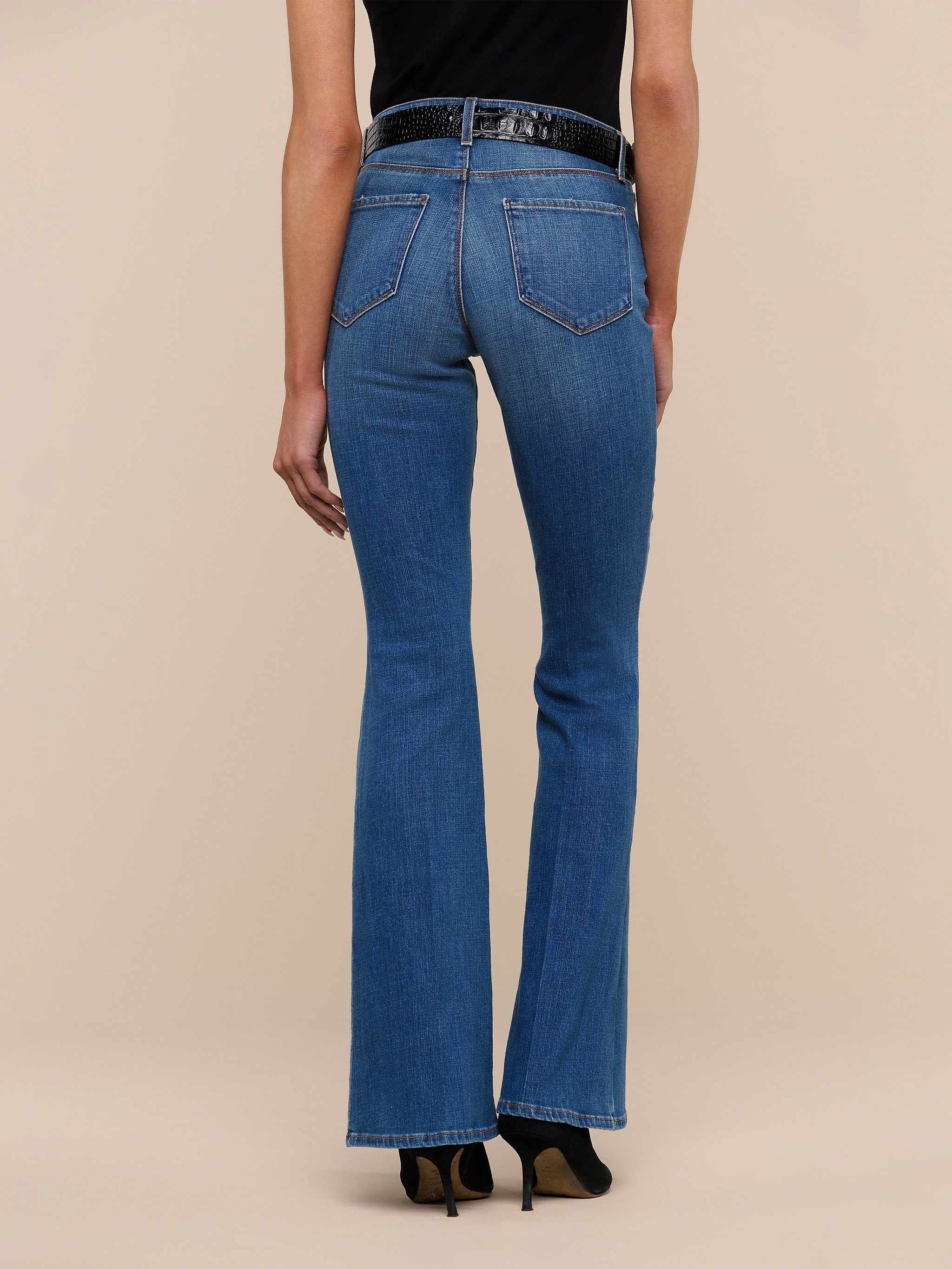 A person stands facing away, wearing the Bell Flare Jean in blue vintage-inspired premium stretch denim, paired with a black top and black heeled shoes against a plain white background.