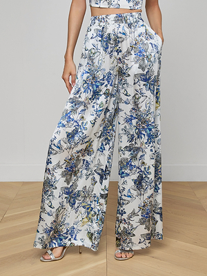A person in Lillian Silk Wide-Leg Pants with a blue, yellow, and grey floral print, styled with a matching crop top and silver heeled sandals, stands on a wooden floor.