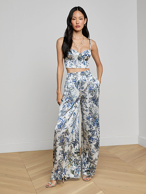 A woman poses indoors in a matching floral crop top and Lillian Silk Wide-Leg Pant set, featuring blue, white, and yellow patterns. She stands on light wood flooring against a plain white wall.