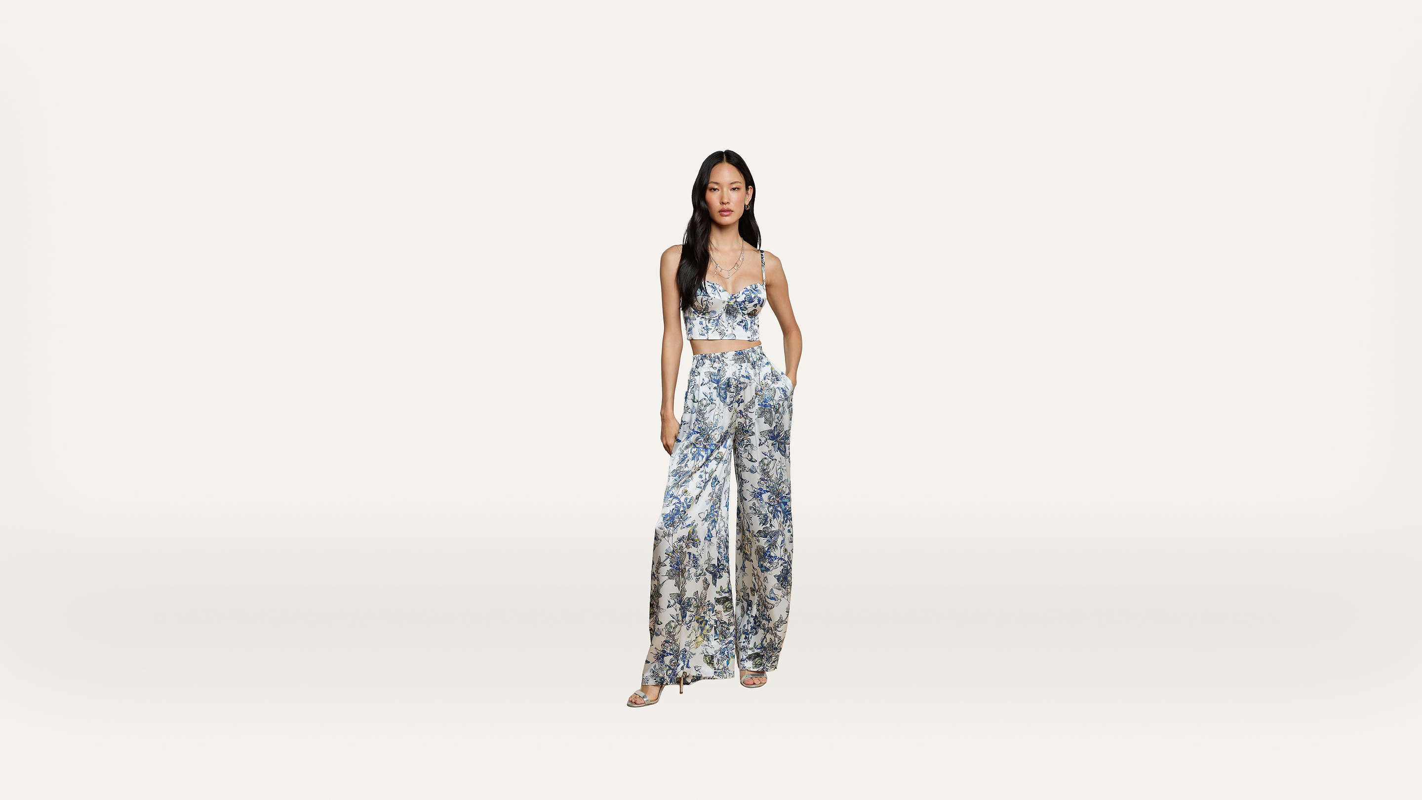 Woman in a blue and white floral patterned crop top and wide-leg pants set, standing against a plain white background, with hands in her pockets and wearing sandals.