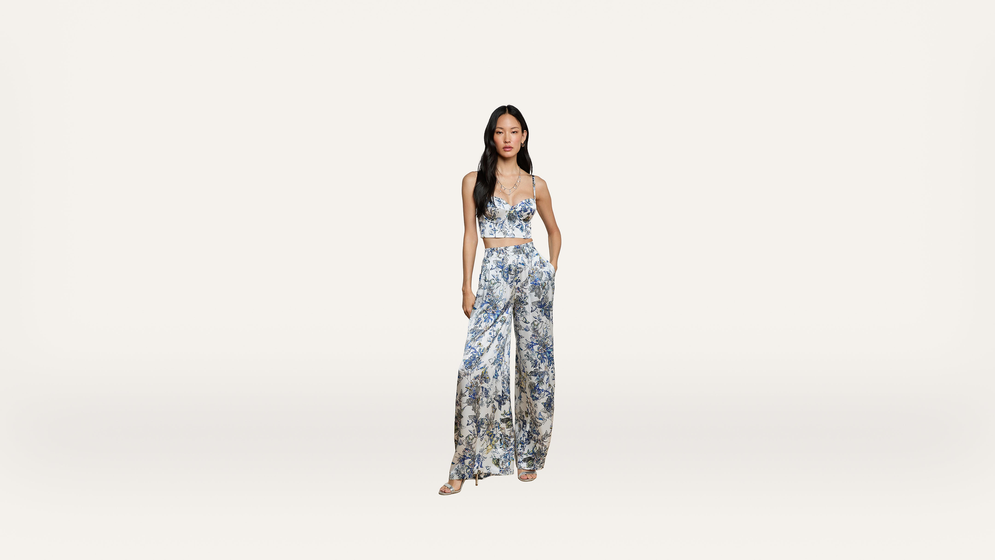 Woman in a blue and white floral patterned crop top and wide-leg pants set, standing against a plain white background, with hands in her pockets and wearing sandals.