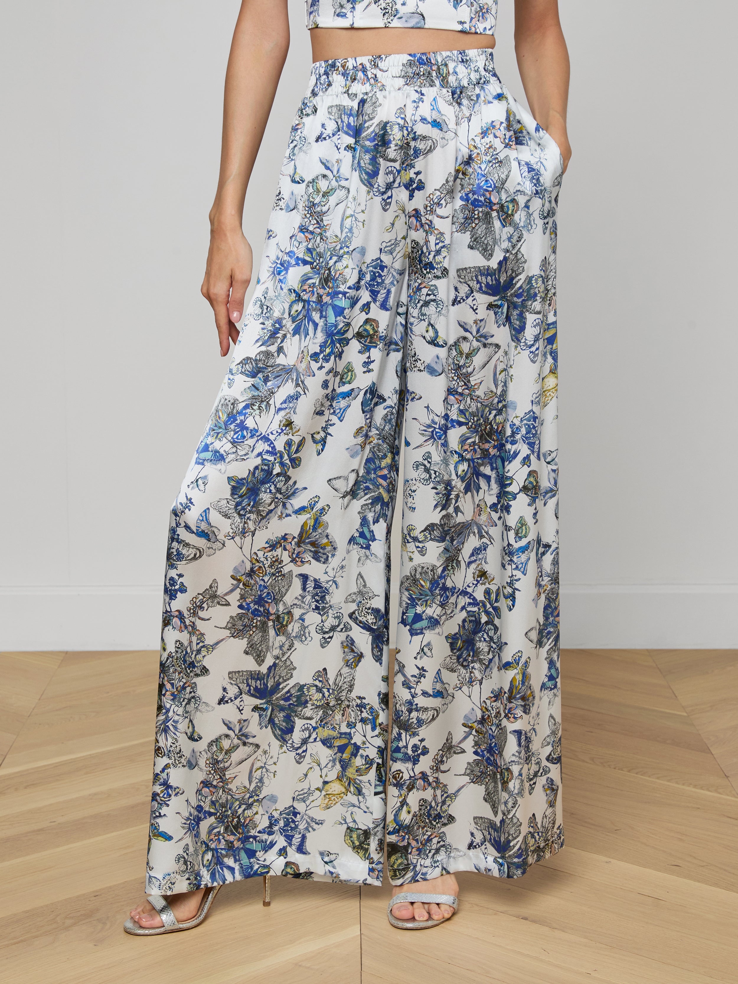 A person wearing Lillian Silk Wide-Leg Pant with a blue, yellow, and green floral butterfly print, a matching crop top, and heeled sandals stands on a light wooden floor.