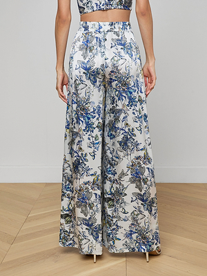 Someone stands indoors on a light wooden floor, facing away, wearing the Lillian Silk Wide-Leg Pant featuring high-waisted, wide-leg styling with a blue, green, and white floral print and an elasticized waistband.