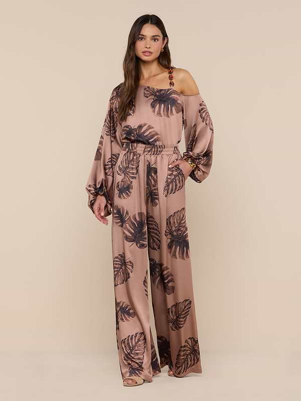 A woman stands against a plain white background, looking at the camera with one hand on her hip, wearing the Lillian Satin Pant in a loose fit with ultra-wide legs and a stylish black palm leaf print.