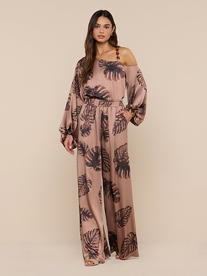 A woman stands against a beige background wearing the Lillian Satin Pant in a black palm leaf print, styled with matching loose, one-shoulder top, open-toed heels, and a bracelet. She has long hair and neutral makeup.