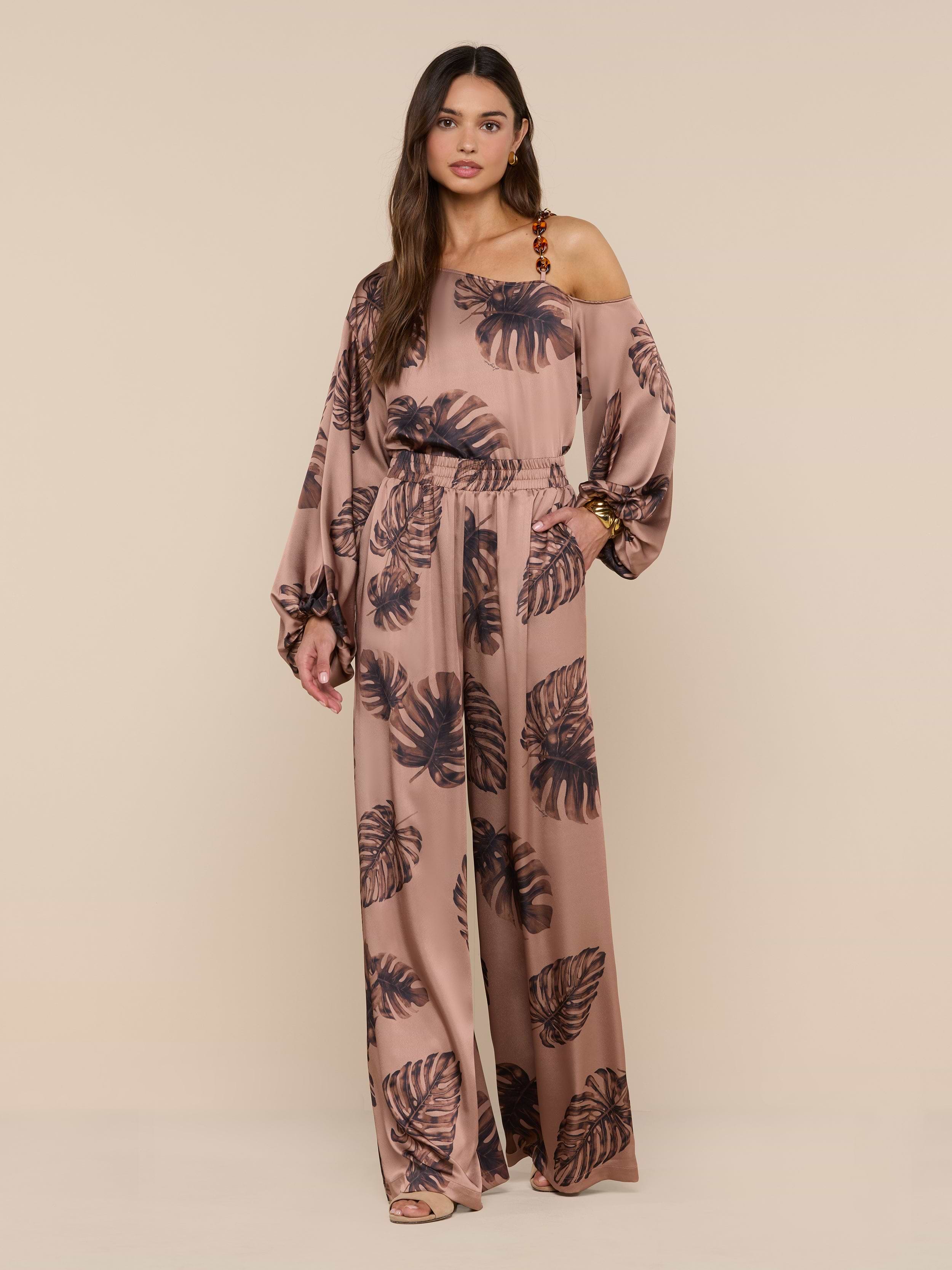 A woman stands against a plain white background, looking at the camera with one hand on her hip, wearing the Lillian Satin Pant in a loose fit with ultra-wide legs and a stylish black palm leaf print.