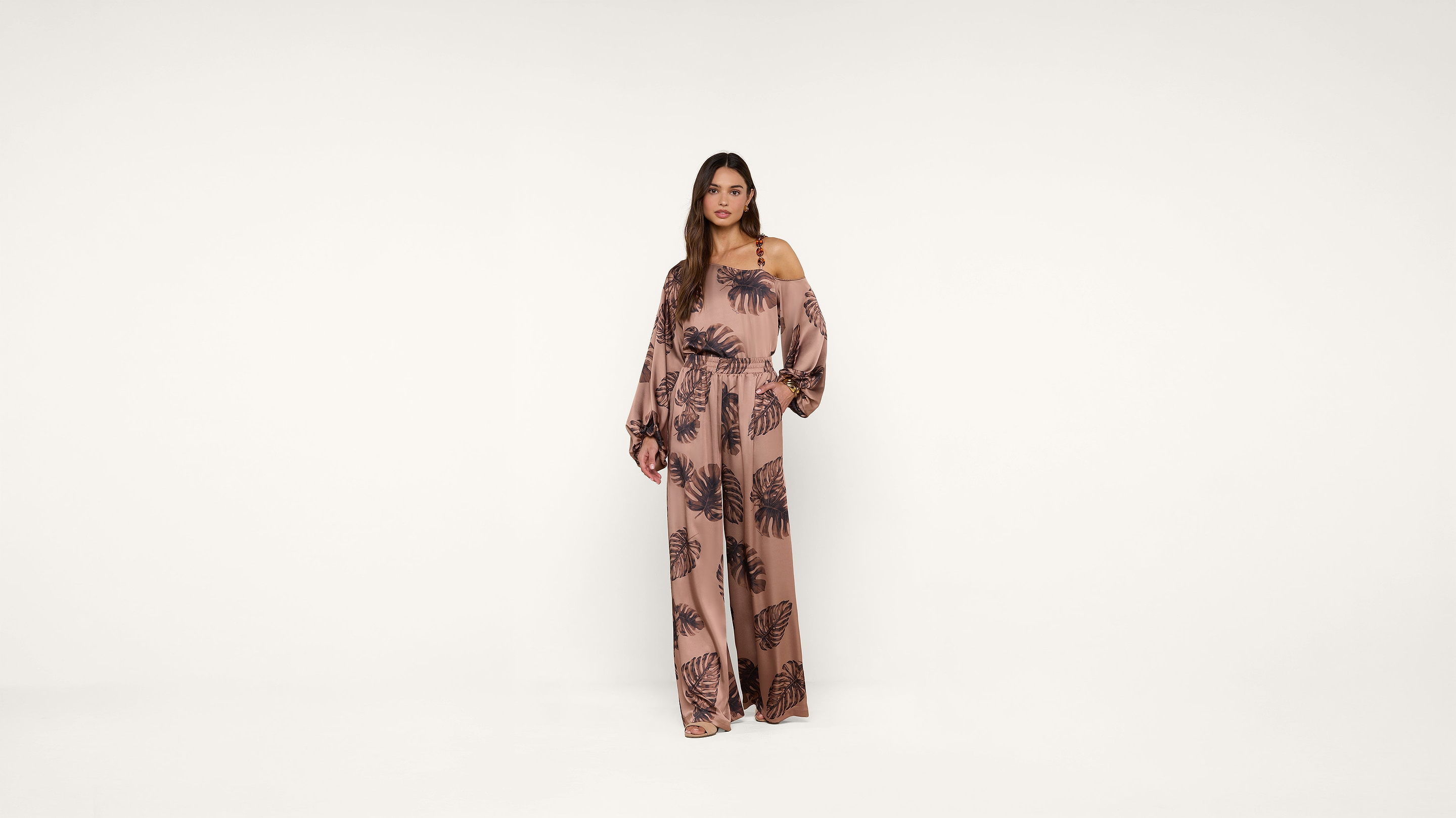 A woman stands against a white background wearing a long, loose-fitting, pink jumpsuit with a dark leaf pattern and wide sleeves.
