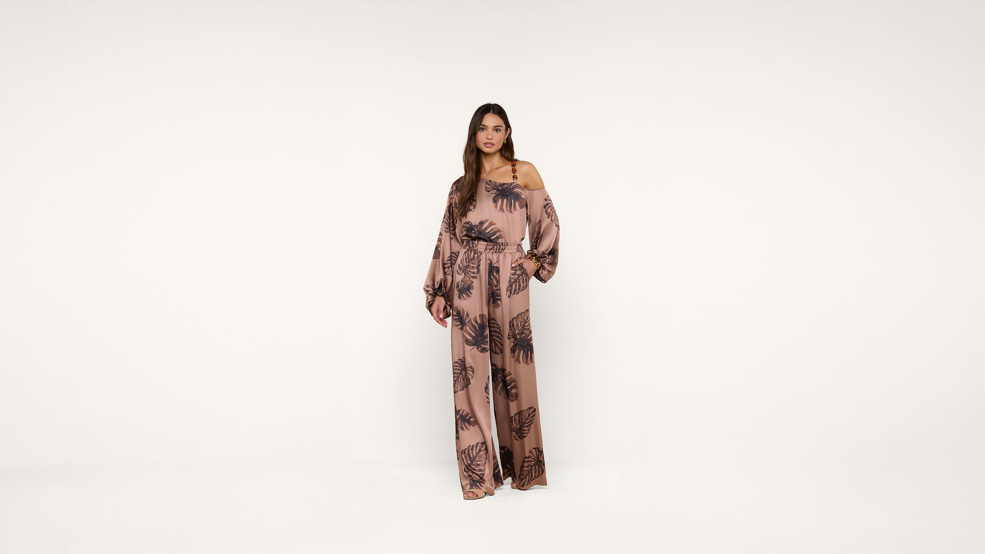 A woman stands against a white background wearing a long, loose-fitting, pink jumpsuit with a dark leaf pattern and wide sleeves.