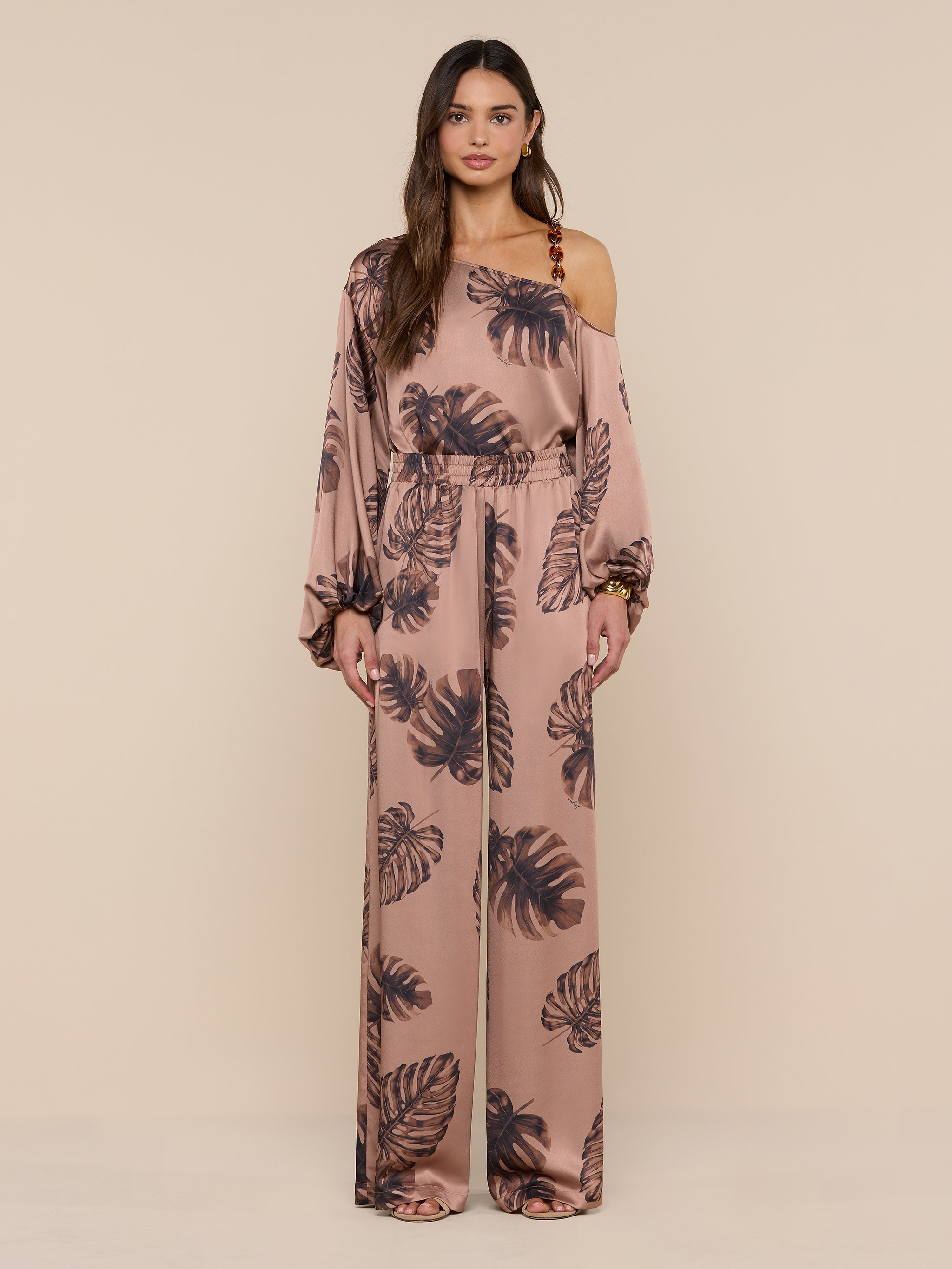 A woman models the Lillian Satin Pant, featuring an ultra-wide silhouette and a dark palm leaf print on pink satin. She stands against a plain white background, looking forward with a neutral expression.