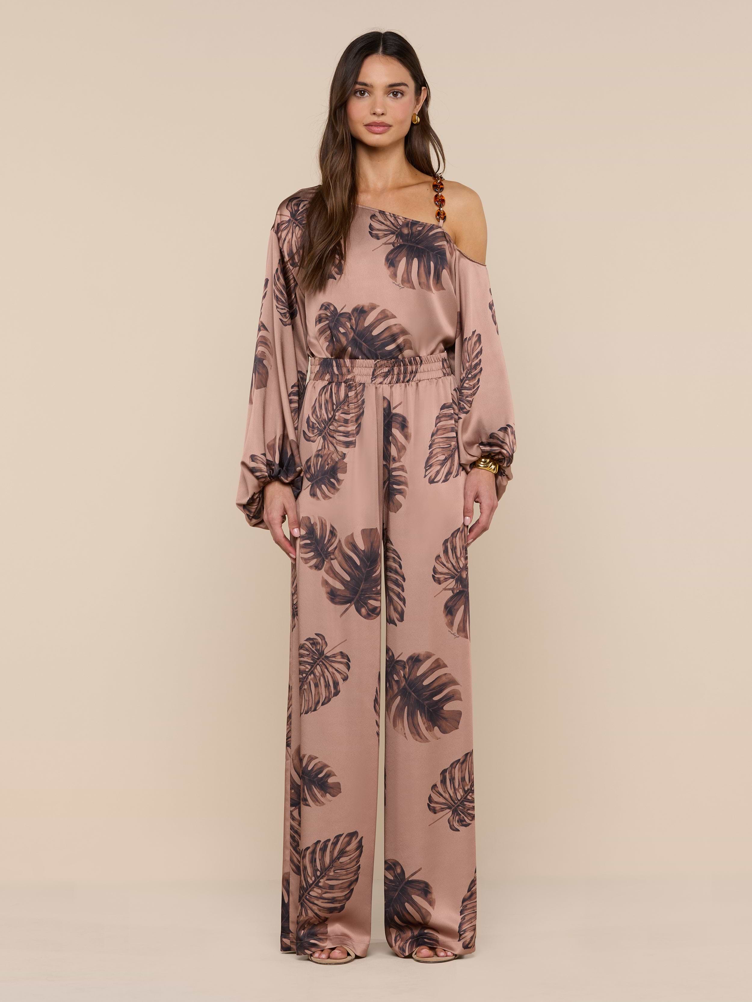 A woman models the Lillian Satin Pant, featuring an ultra-wide silhouette and a dark palm leaf print on pink satin. She stands against a plain white background, looking forward with a neutral expression.