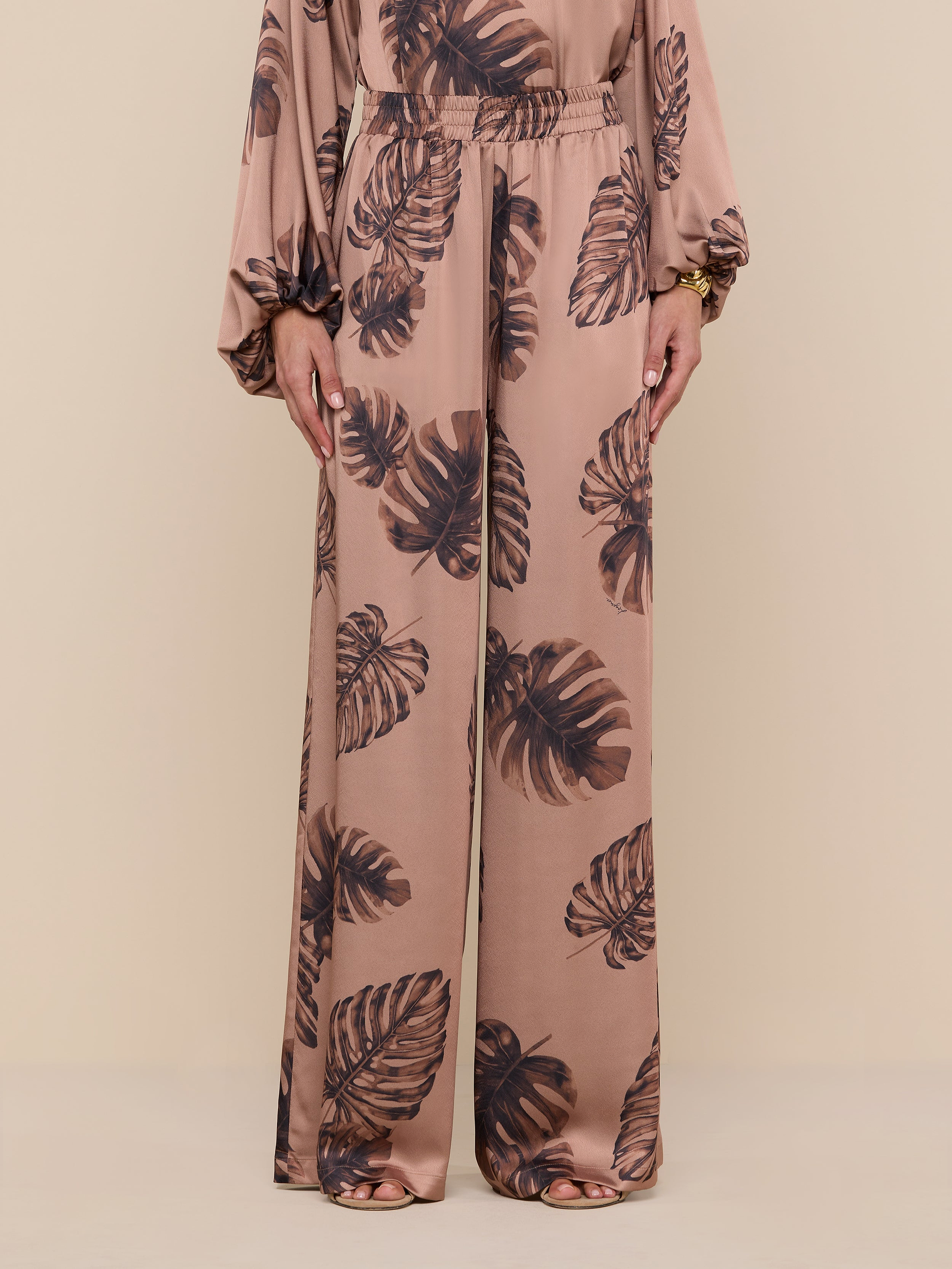 A person wearing the Lillian Satin Pant, featuring an ultra-wide leg and light brown satin fabric with a dark brown palm leaf print, stands against a plain white background; only their lower body is visible.