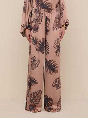 A person models the Lillian Satin Pant—ultra-wide, brown with a palm leaf pattern—in lustrous satin against a beige backdrop. Visible from shoulders down, their hands rest at their sides.