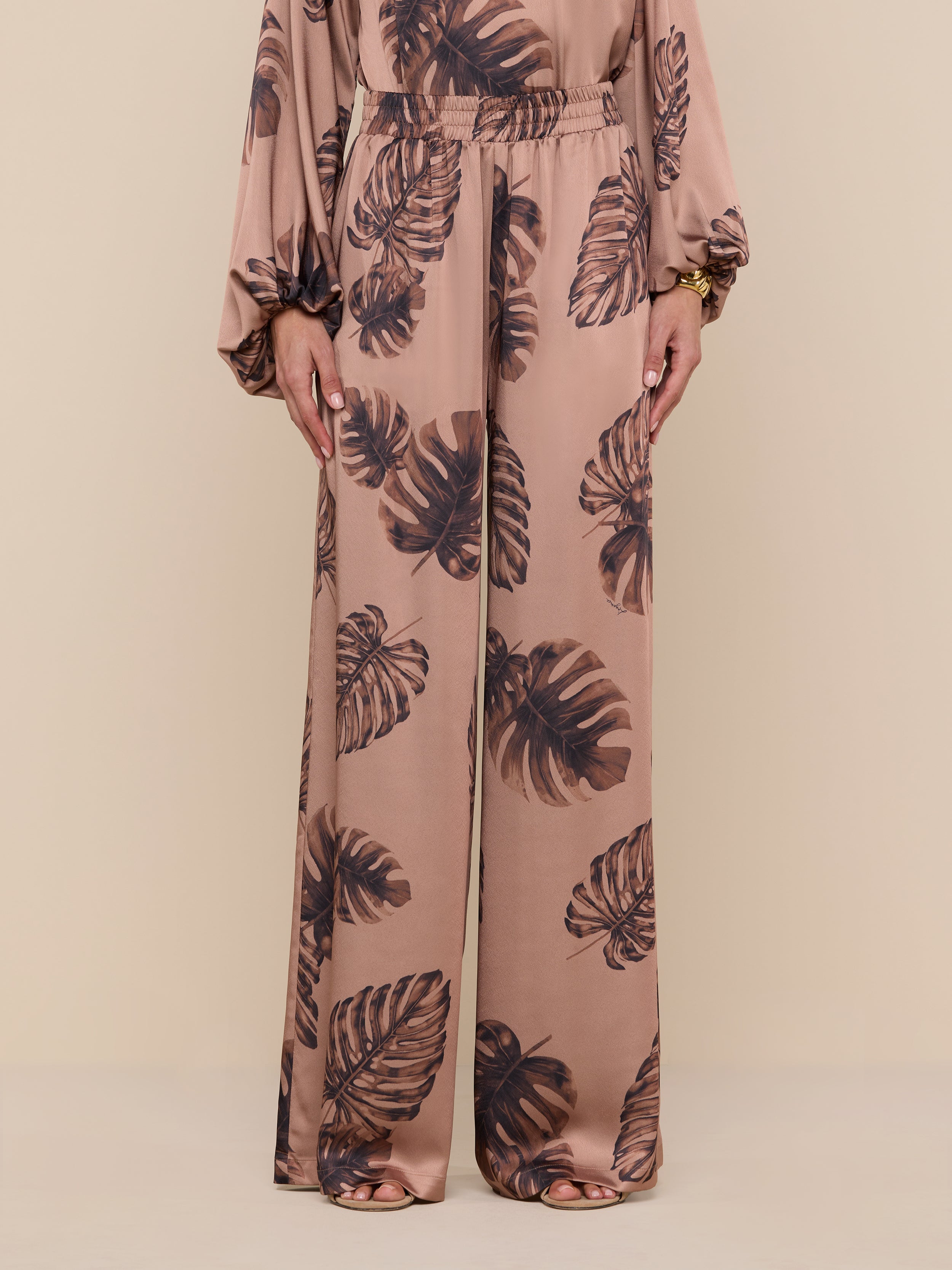 A person wearing the Lillian Satin Pant, featuring an ultra-wide leg and light brown satin fabric with a dark brown palm leaf print, stands against a plain white background; only their lower body is visible.