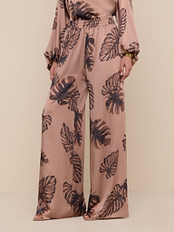 A person stands against a plain background, showcasing only the lower half of their outfit and wearing the Lillian Satin Pant featuring an ultra-wide silhouette with a brown palm leaf print.