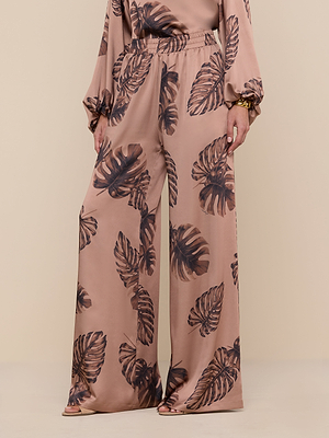A person models the Lillian Satin Pant—ultra-wide, high-waisted, light brown satin pants with a dark brown palm leaf print—paired with a matching long-sleeve top and open-toe shoes against a beige background.