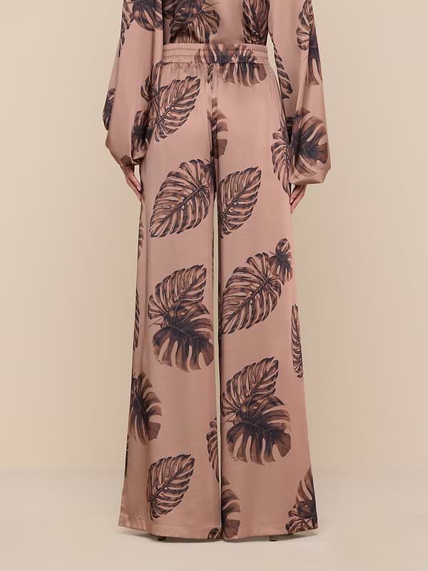 A person models the Lillian Satin Pant in an ultra-wide, high-waisted silhouette, featuring a large dark palm leaf print on a brown background. The matching long-sleeve top is partially visible. Only the lower body and hands are shown against a plain white backdrop.