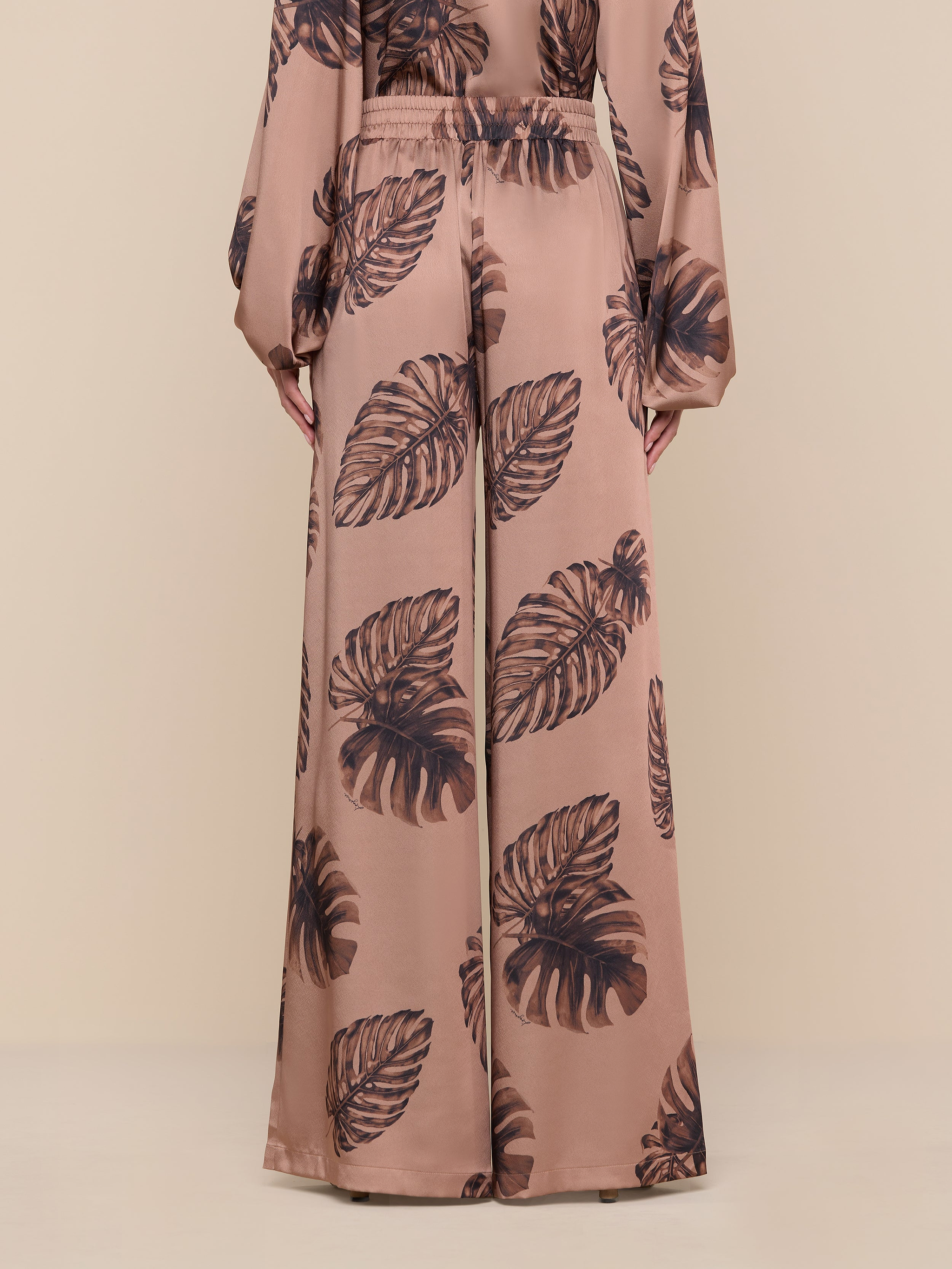 A person models the Lillian Satin Pant in an ultra-wide, high-waisted silhouette, featuring a large dark palm leaf print on a brown background. The matching long-sleeve top is partially visible. Only the lower body and hands are shown against a plain white backdrop.
