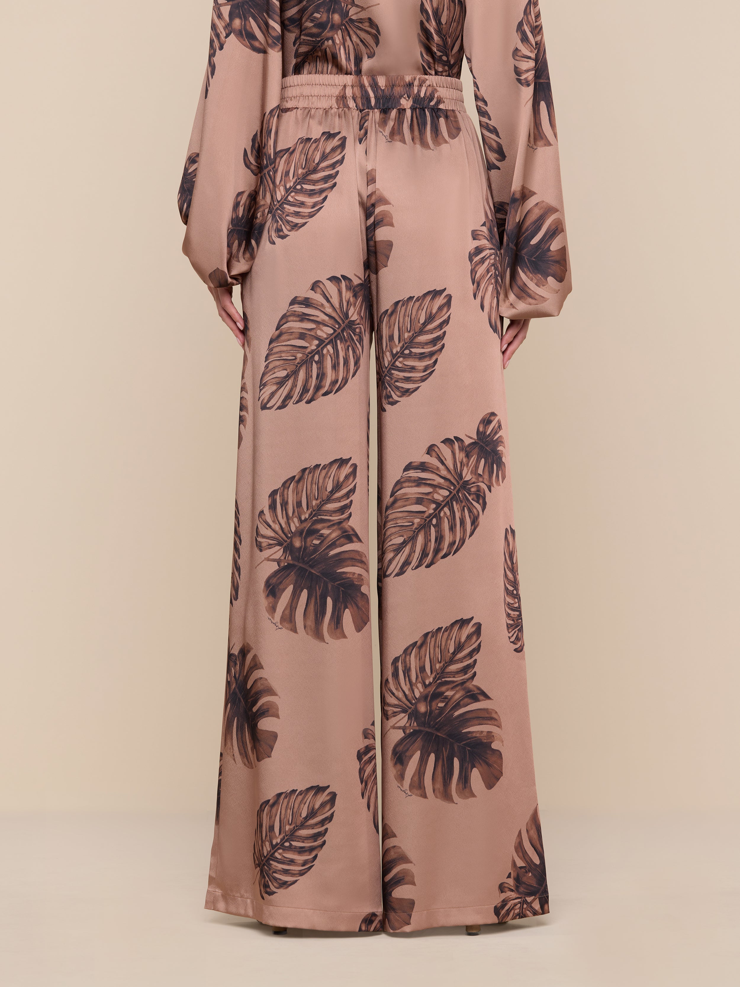A person models the Lillian Satin Pant in an ultra-wide, high-waisted silhouette, featuring a large dark palm leaf print on a brown background. The matching long-sleeve top is partially visible. Only the lower body and hands are shown against a plain white backdrop.