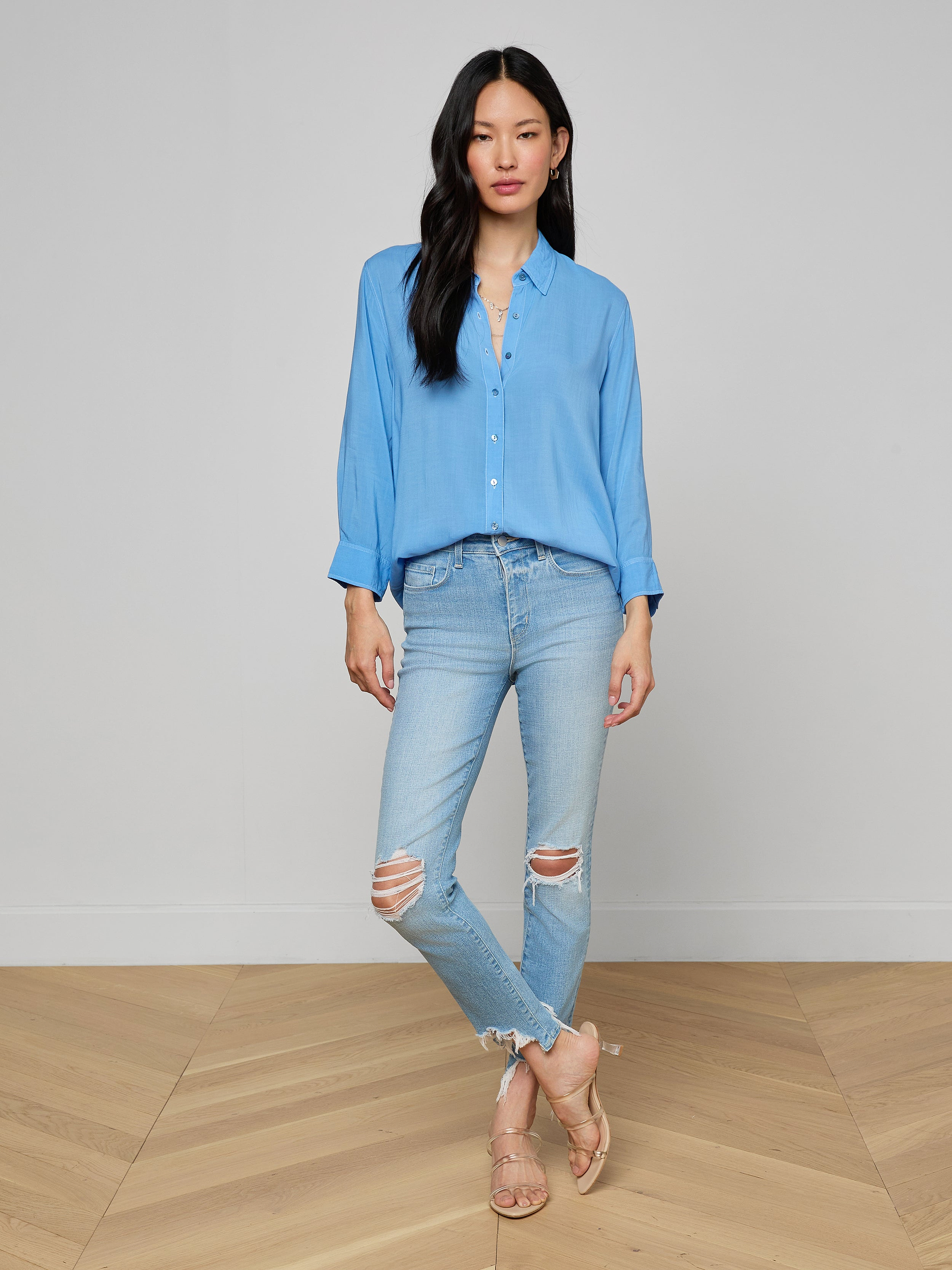 A woman stands against a plain background, wearing a light blue button-up shirt paired with the High Line Distressed Skinny Jean in a vintage wash with rips, and beige heeled sandals. She has long black hair and her arms are relaxed at her sides.