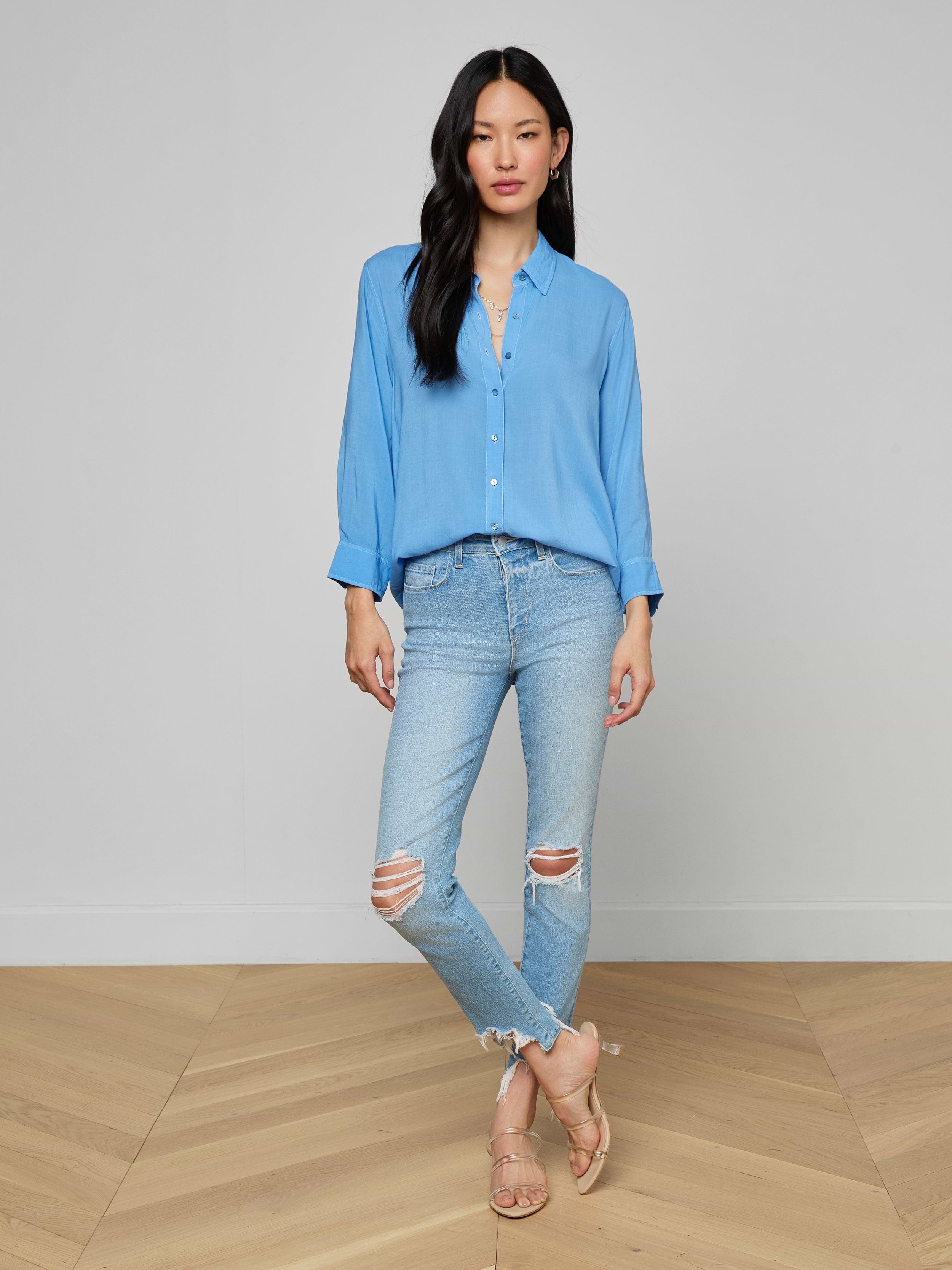 A woman stands against a plain background, wearing a light blue button-up shirt paired with the High Line Distressed Skinny Jean in a vintage wash with rips, and beige heeled sandals. She has long black hair and her arms are relaxed at her sides.