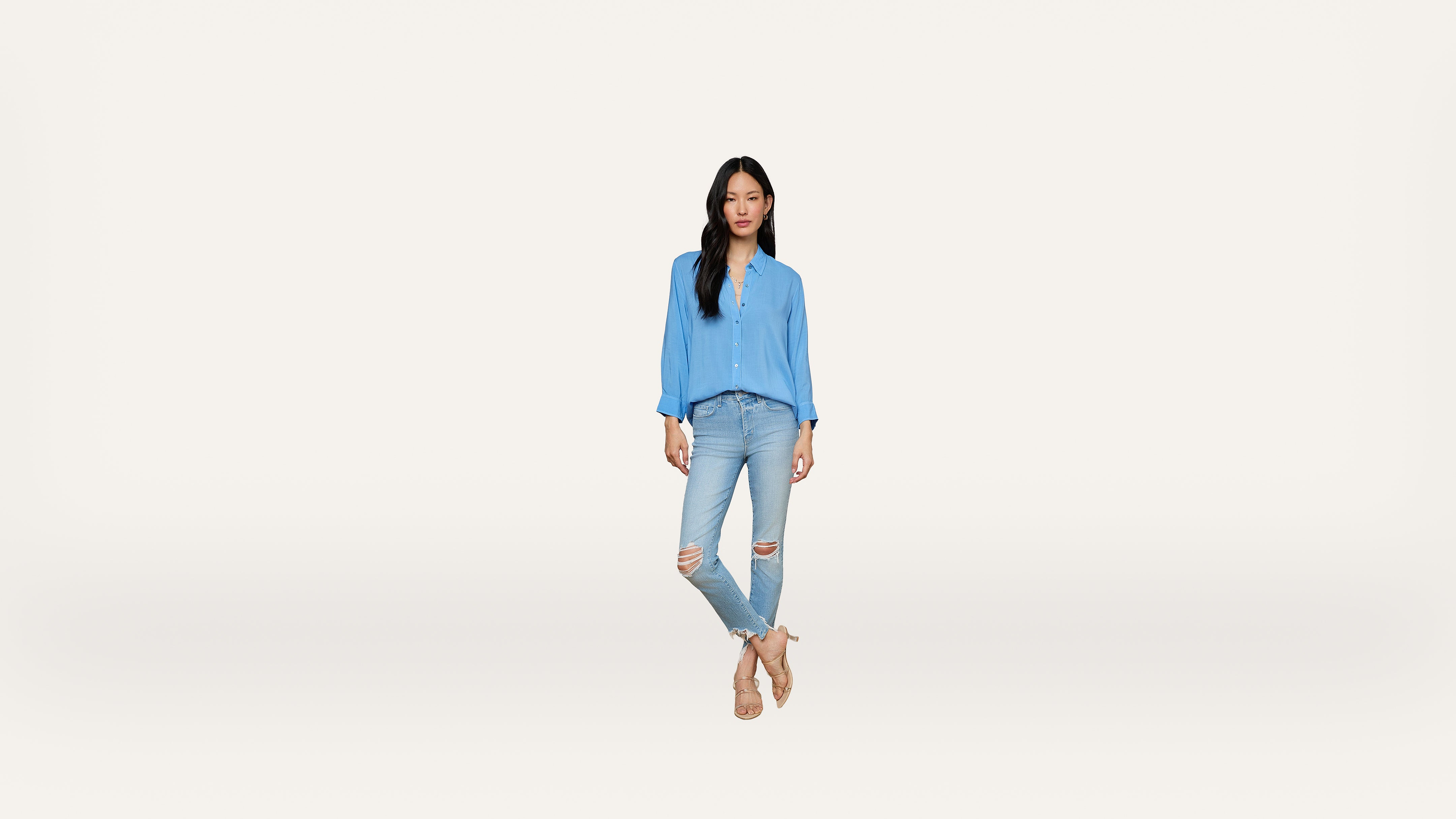 Woman standing against a plain white background, wearing a light blue blouse, ripped light blue jeans, and beige sandals. She has long dark hair and appears relaxed with her hands in her pockets.