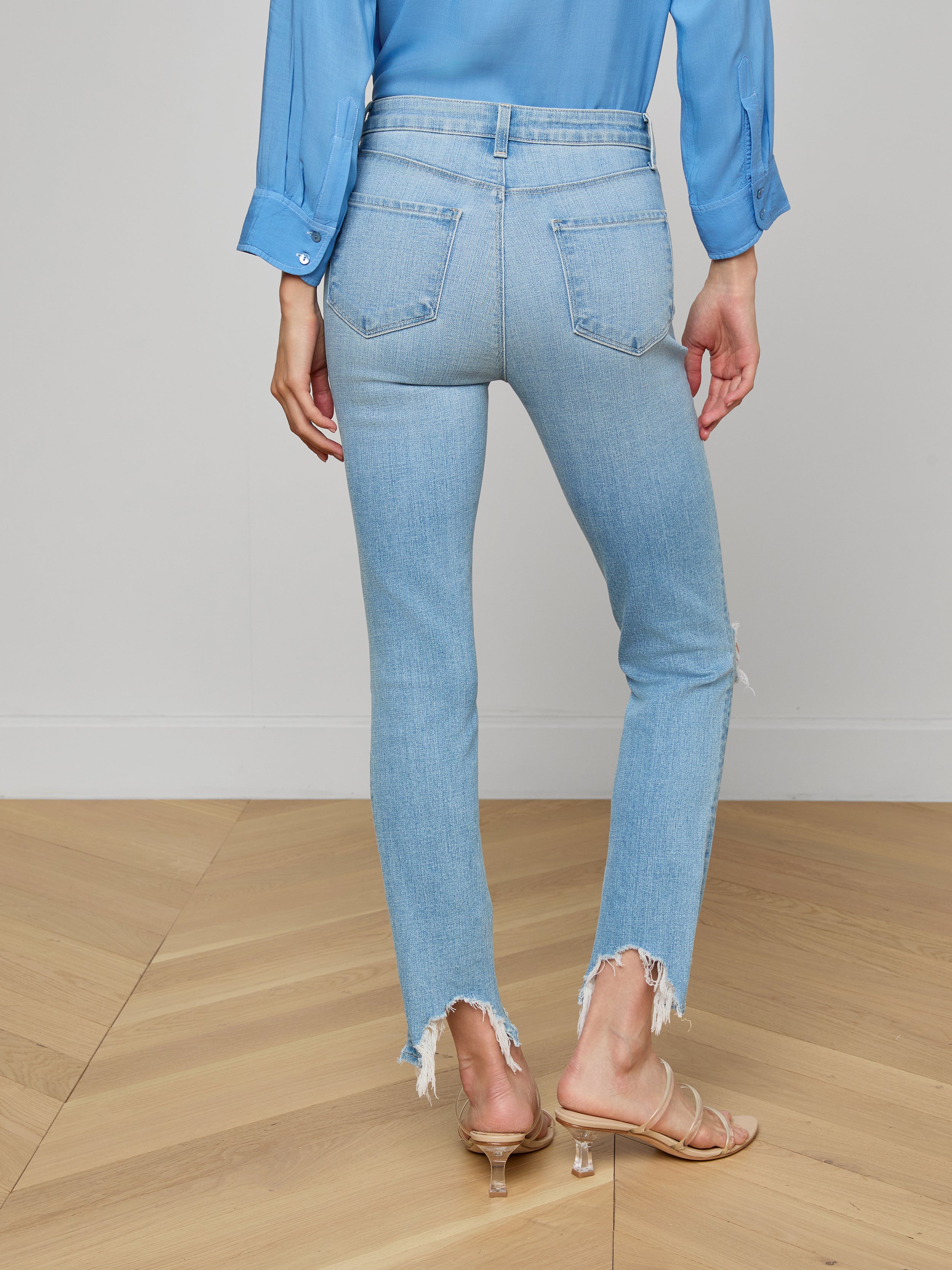 A woman stands with her back to the camera against a plain white background, wearing the High Line Distressed Skinny Jean in a light blue vintage wash with frayed hems, paired with a blue long-sleeve shirt and beige heeled sandals.