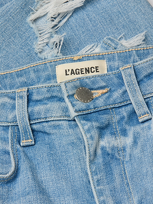 Close-up of the High Line Distressed Skinny Jean in light blue vintage wash, showing frayed rip detail, a white LAGENCE label above the waistband, and a metal button.