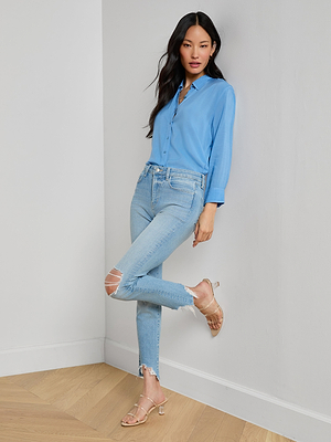 A woman stands indoors against a light wall, wearing a light blue button-up shirt and High Line Distressed Skinny Jeans with a vintage wash, paired with beige sandals. One leg is crossed over the other and her hands rest casually by her sides.